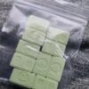 Pale green, square tablets with embossed designs inside a clear resealable plastic bag on a textured fabric surface.